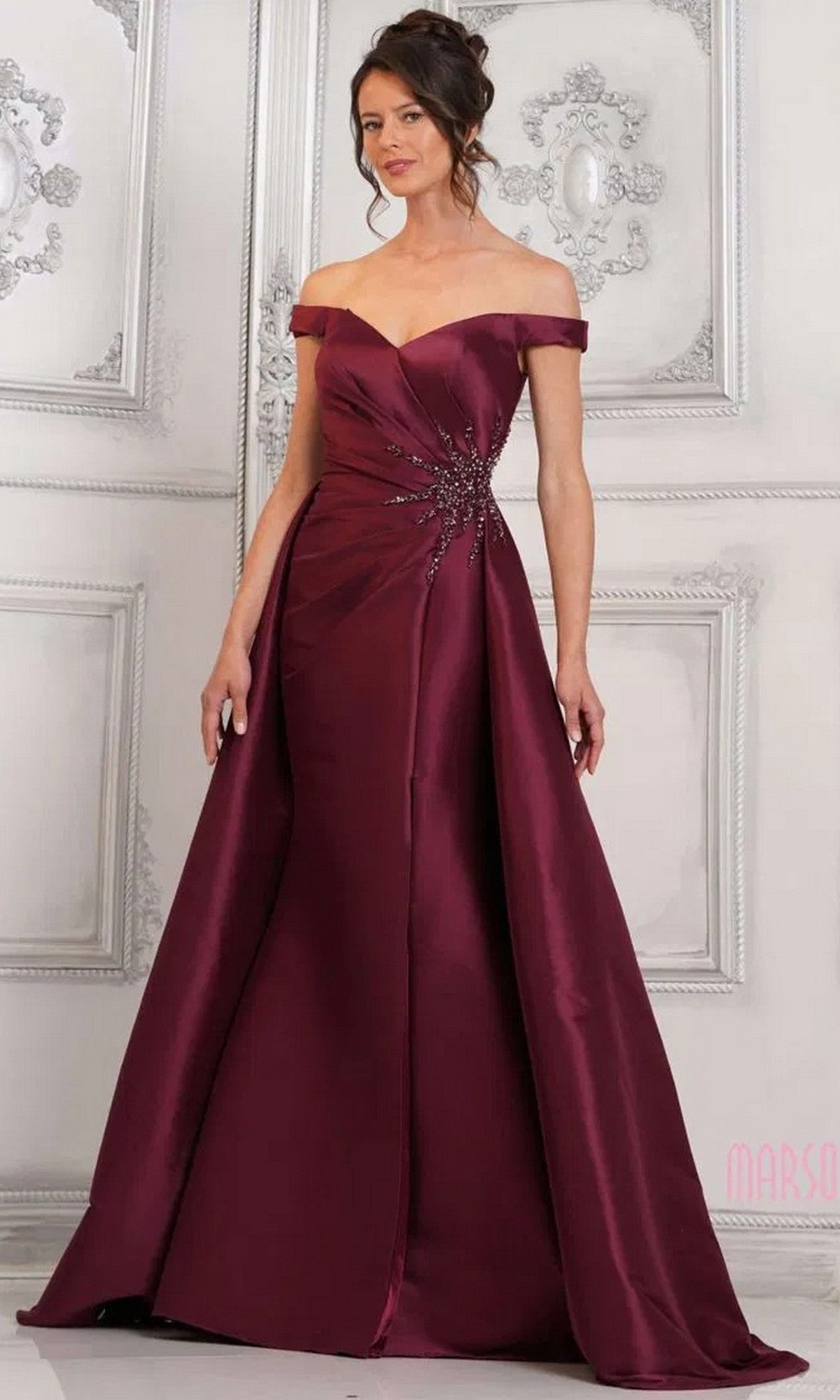 A woman stands before an ornate white wall wearing the Marsoni MV1310 long formal dress in wine, featuring off-the-shoulder styling, a floor-length mermaid silhouette, and embellished waist details.